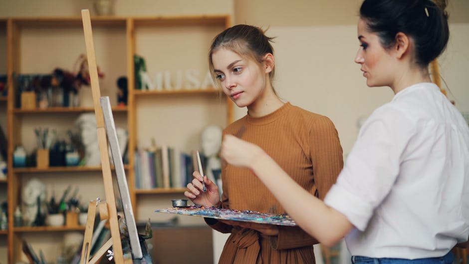 Female art instructor teaching a young woman painting techniques in a cozy studio.