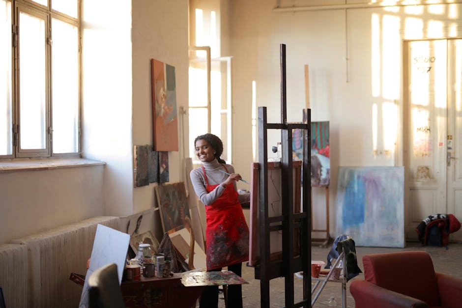 Smiling black lady in apron and casual clothes standing near easel in creative workshop and painting pictures on canvas while looking at camera